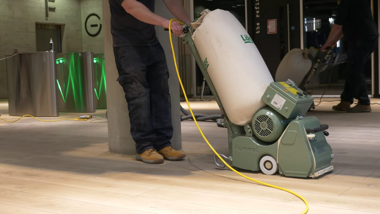 Commercial Floor Sanding in Stamford Hill, with a technician using a Hummel belt sander on a light oak hardwood reception floor to create a smooth, even surface.