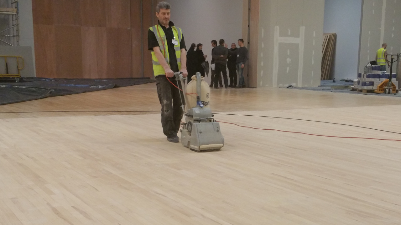 Technician carrying out Commercial Floor Sanding in Stamford Hill on a large maple strip hardwood sports hall floor using a professional belt sander.